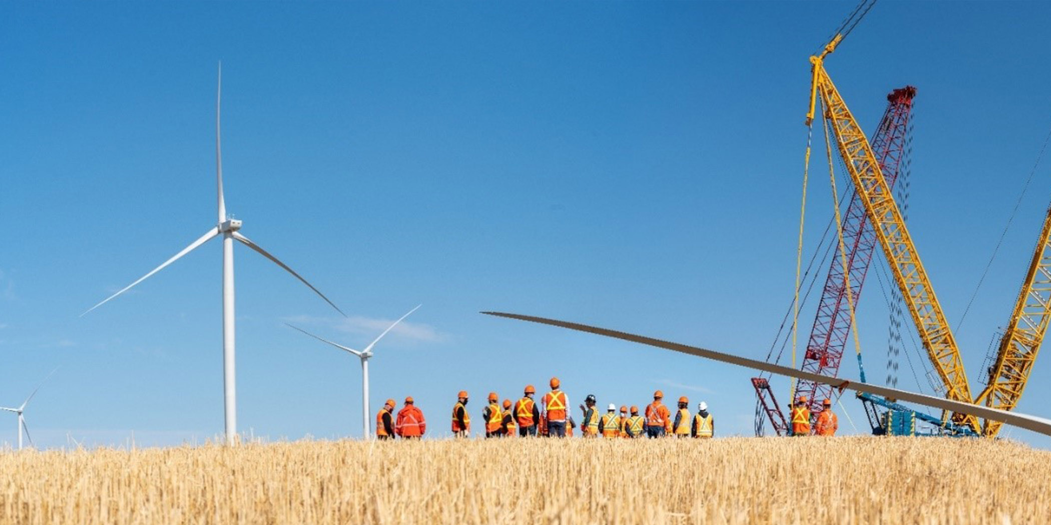 2 Wind turbines in the background with a group of workers dressed in orange around equipment