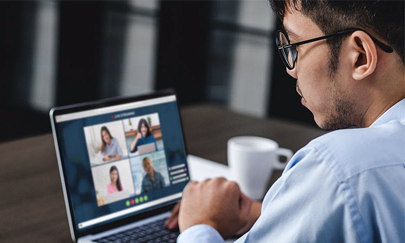 A male wearing a blue shirt looking at a screen - while video chatting with 4 other people.