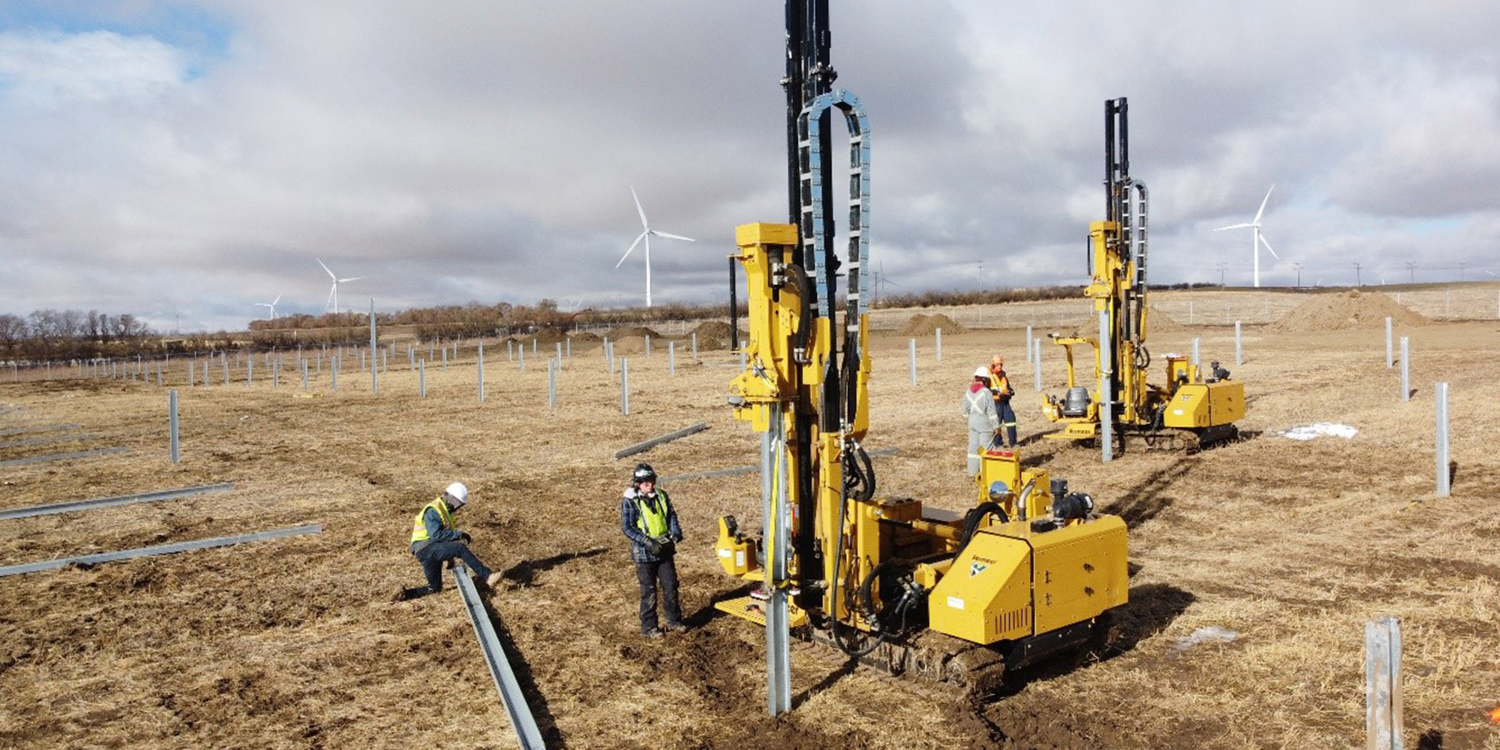 2 pile machines in a field hammering in metal piles with workers nearby