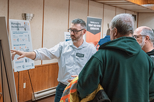 A male pointing to a post board that 2 people are looking at.